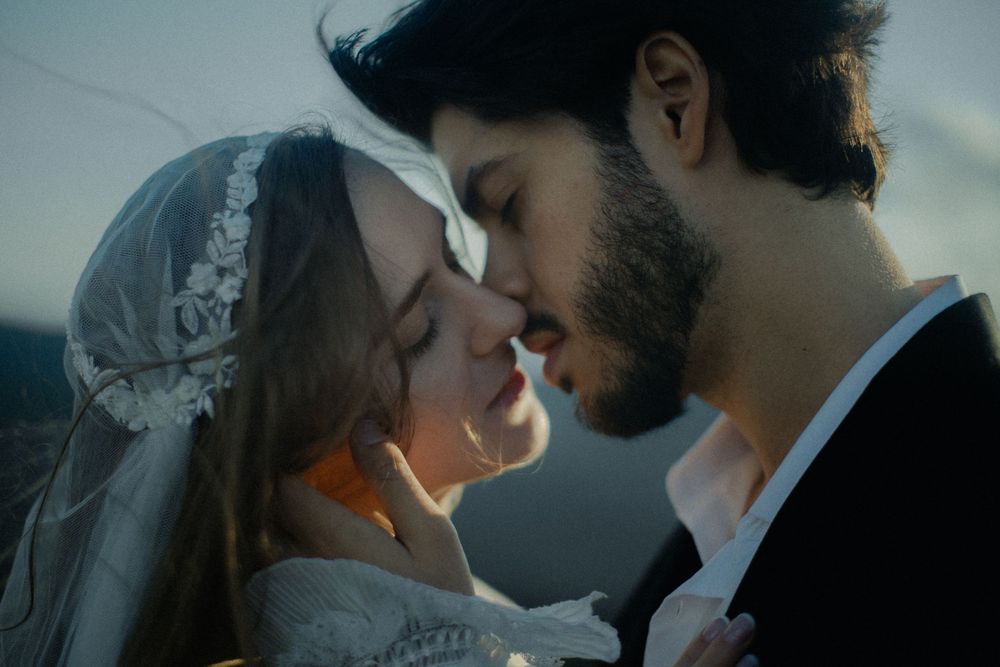 A bride and groom share a romantic almost-kiss moment outdoors, wearing wedding attire in soft evening light.