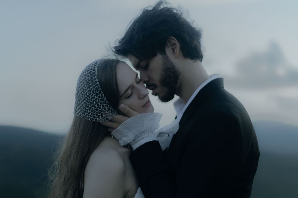 A romantic couple in vintage wedding attire share an intimate moment against a moody, misty landscape.