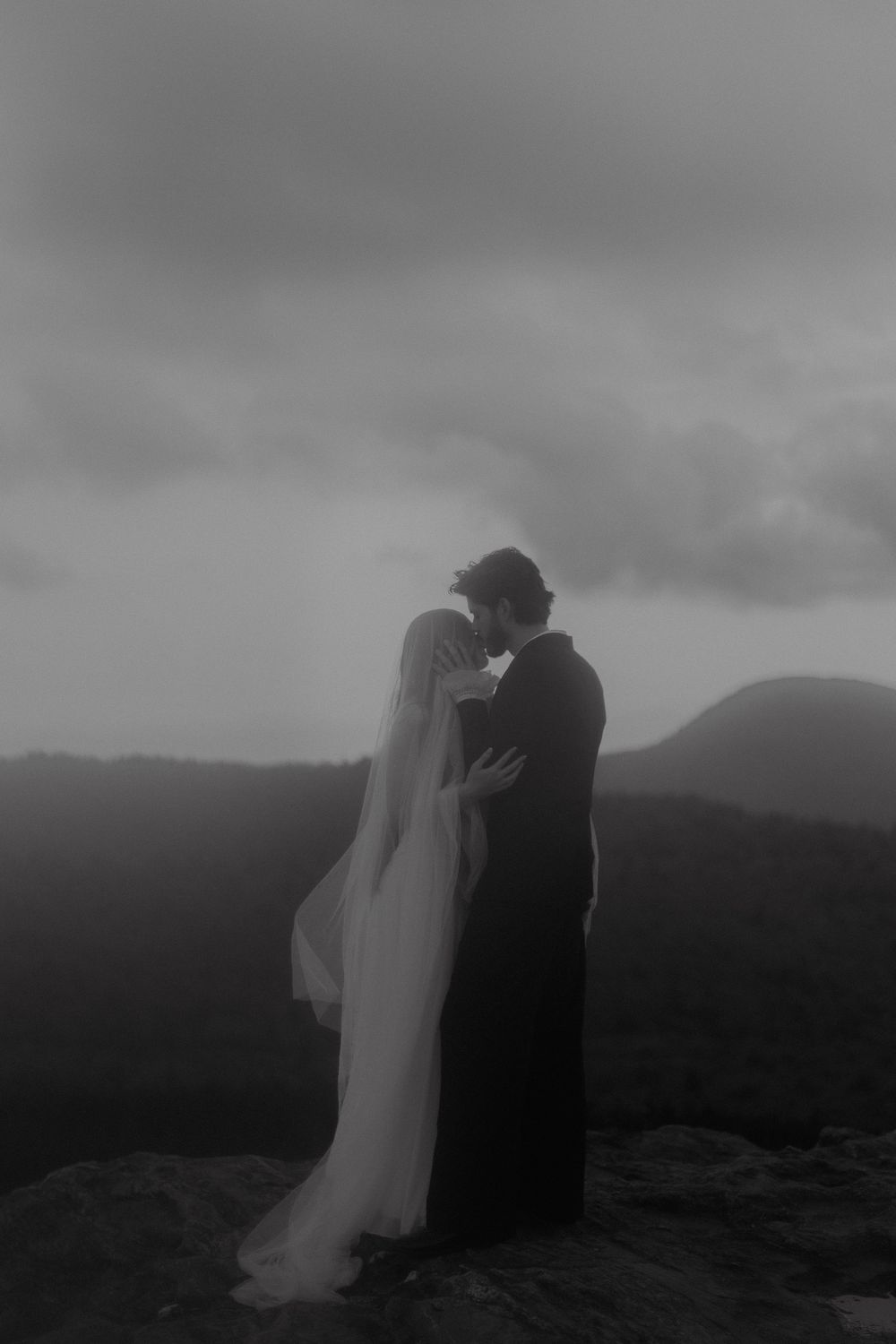 Black and white photo of a couple sharing a romantic kiss on a misty mountain summit, bride in flowing veil.