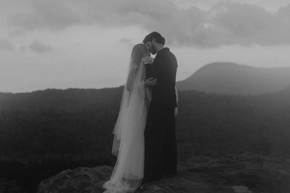 Black and white photo of a bride and groom kissing on a rocky mountain summit with misty hills in the background.