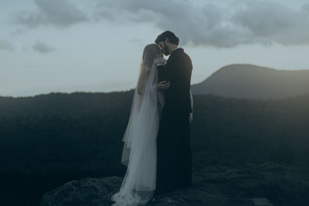 A bride and groom share a romantic kiss on a rocky mountaintop at dusk, surrounded by misty mountain views.