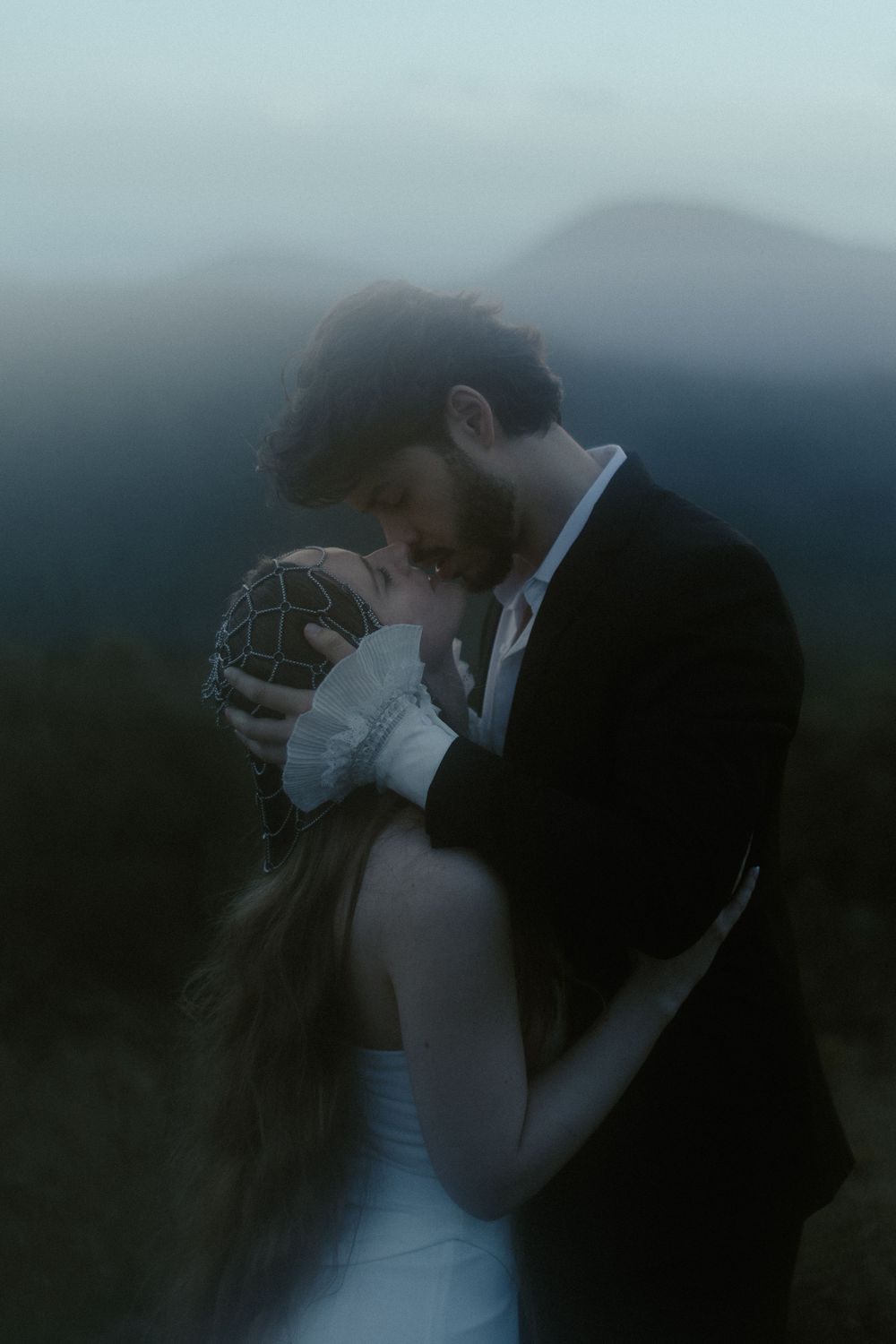 A couple shares an intimate embrace at dusk with misty mountains in the background, the bride in white and groom in black.