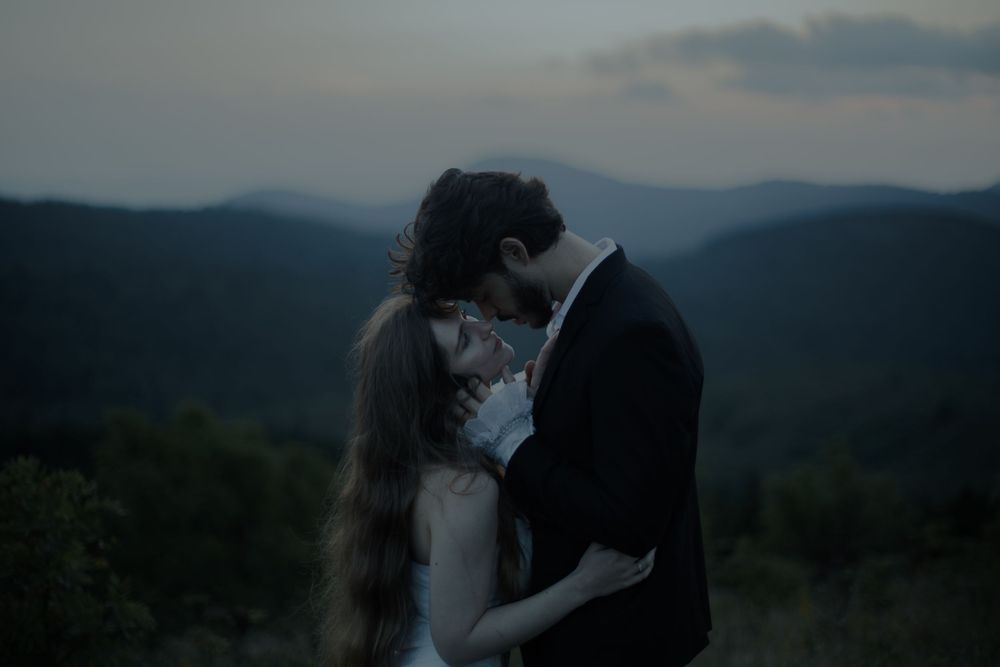A romantic couple embraces at dusk with misty mountain forests in the background, bride in white gown.