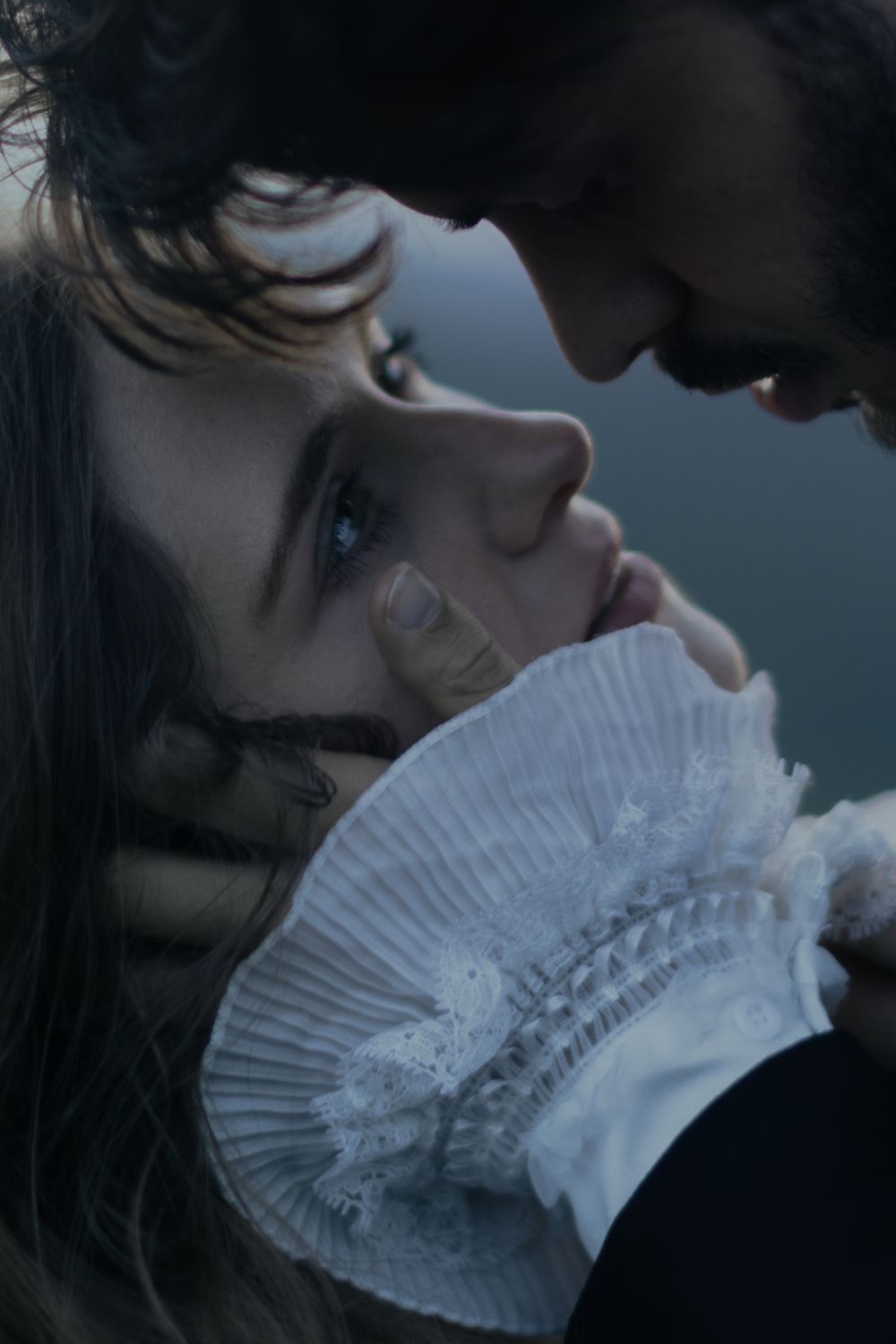 A romantic close-up of a couple in period costume, faces nearly touching, with a white ruffled lace cuff visible.