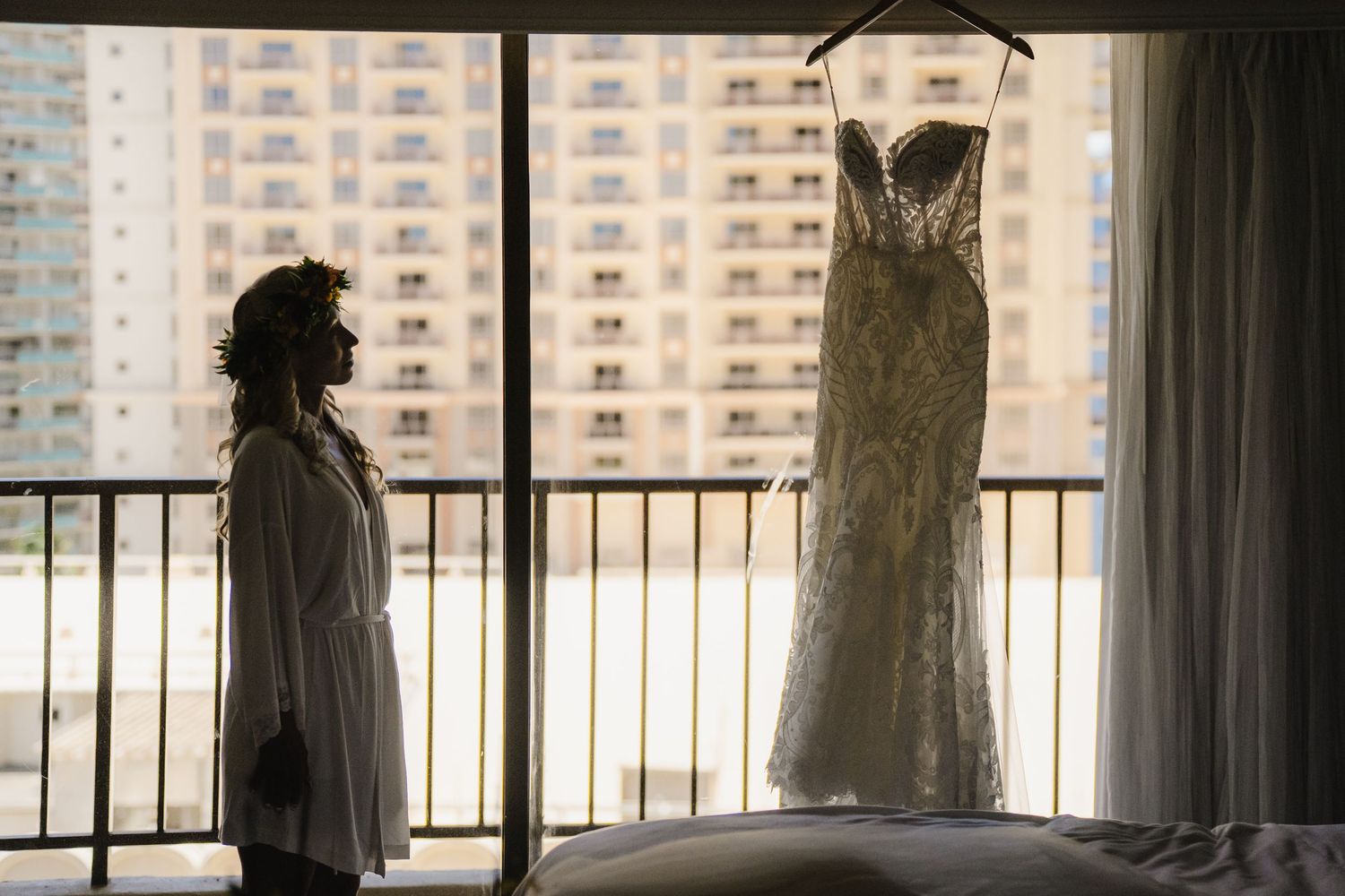 Bride silhouetted beside elegant lace wedding dress hanging on balcony window with city skyline backdrop.