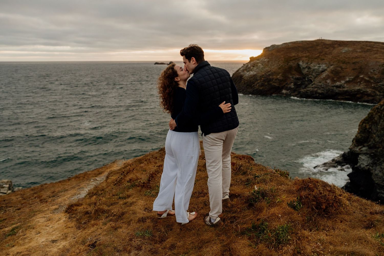Couple sharing a romantic kiss on a coastal cliff at sunset with dramatic ocean views and moody skies.