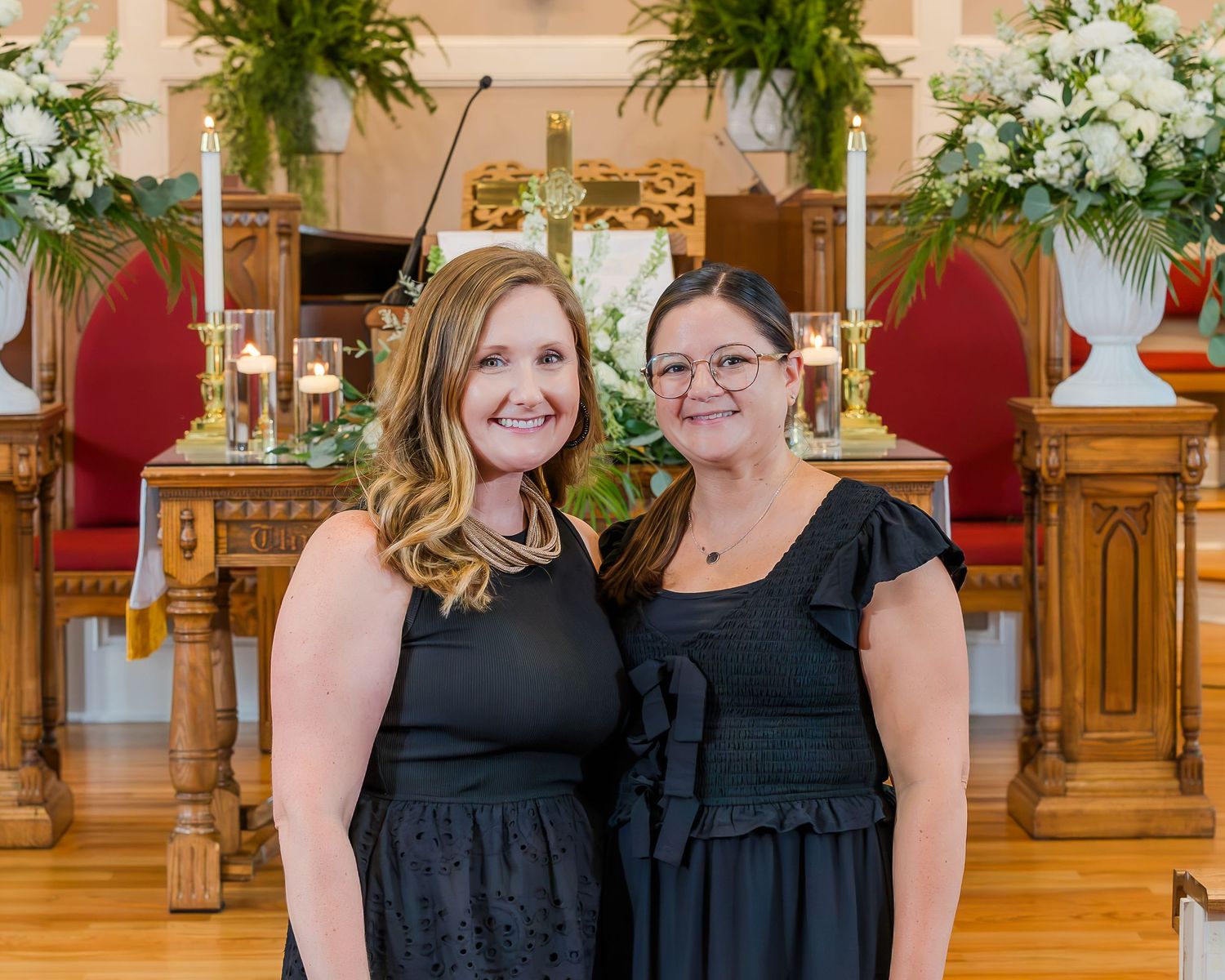 Two women in black dresses smiling together inside a decorated church with floral arrangements.