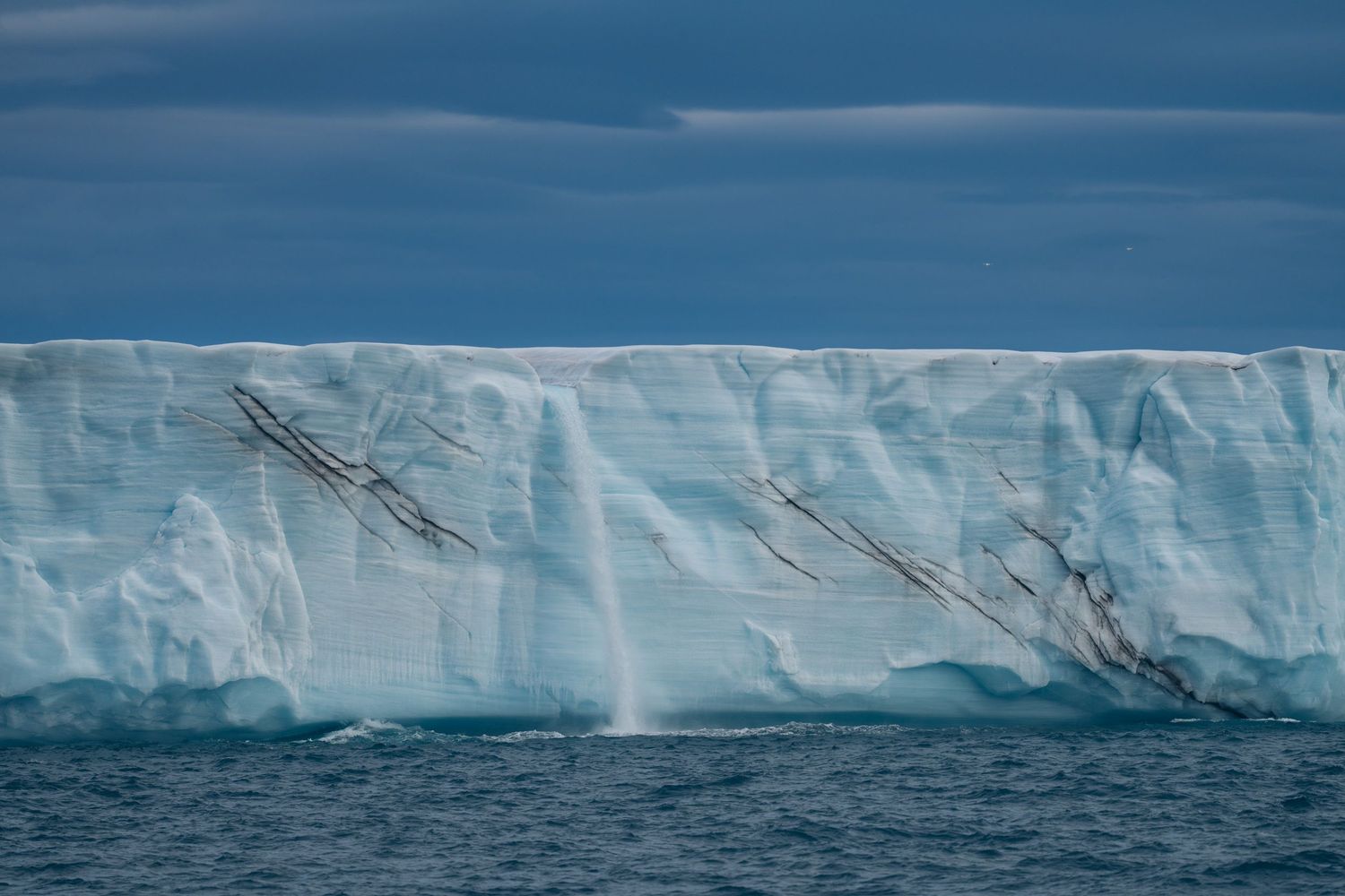 Veines de moraine sombre striant le front de glace de Bråsvellbreen, cascade au centre