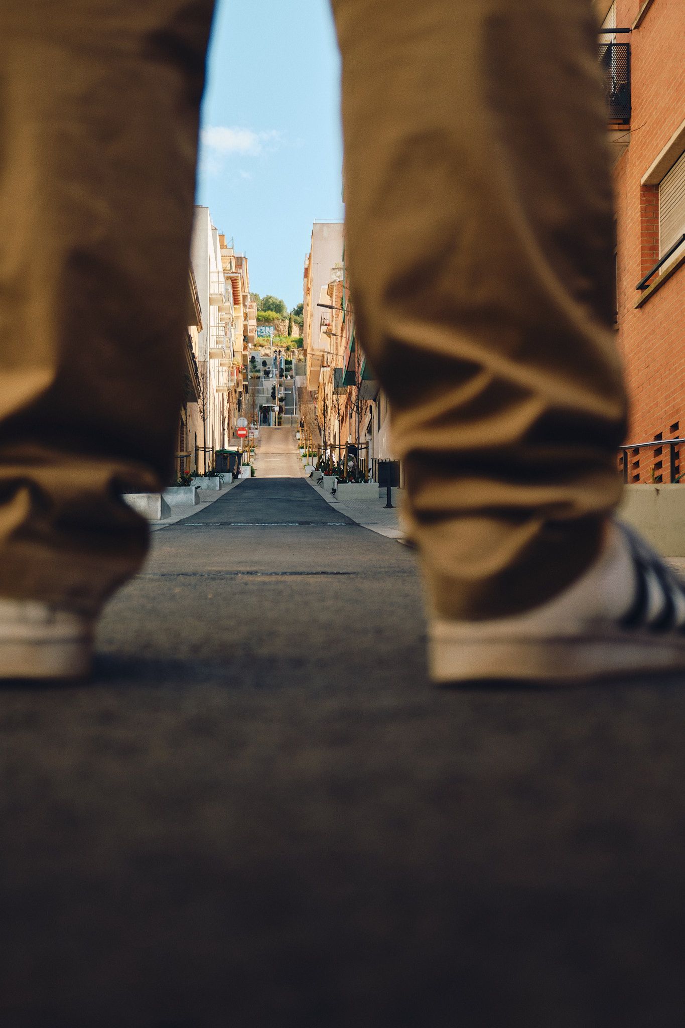Low-angle street view between two people wearing tan pants on a narrow urban alley with buildings and blue sky ahead.