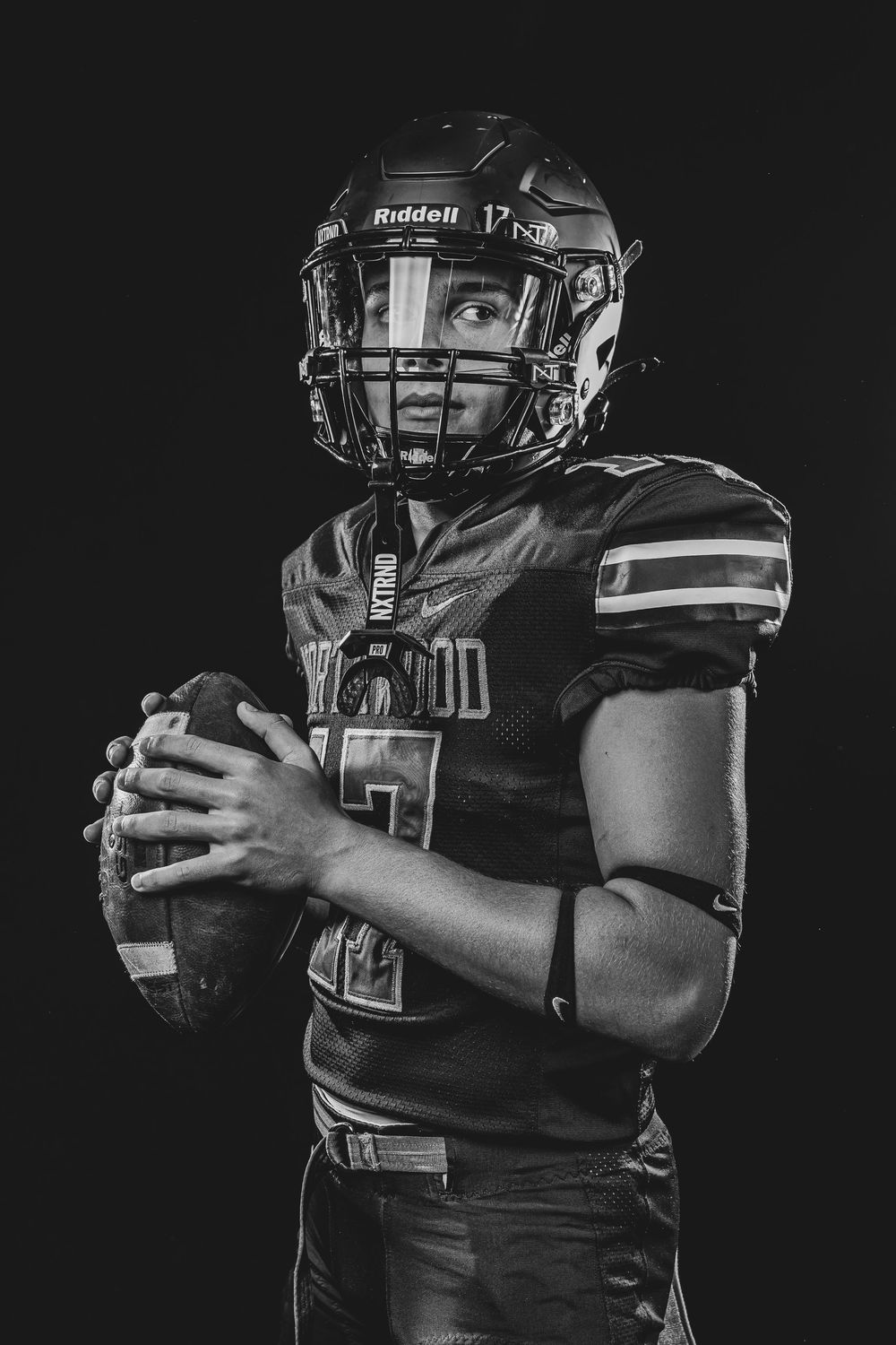 Black and white portrait of a football player in Riddell helmet holding a football against a dark background.