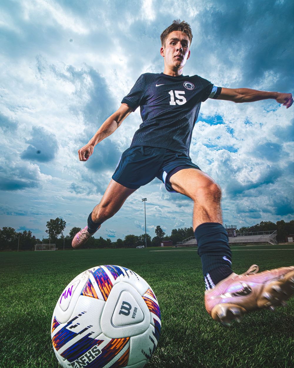 Soccer player in navy #15 Penn State jersey kicking a ball on green field under dramatic cloudy sky.