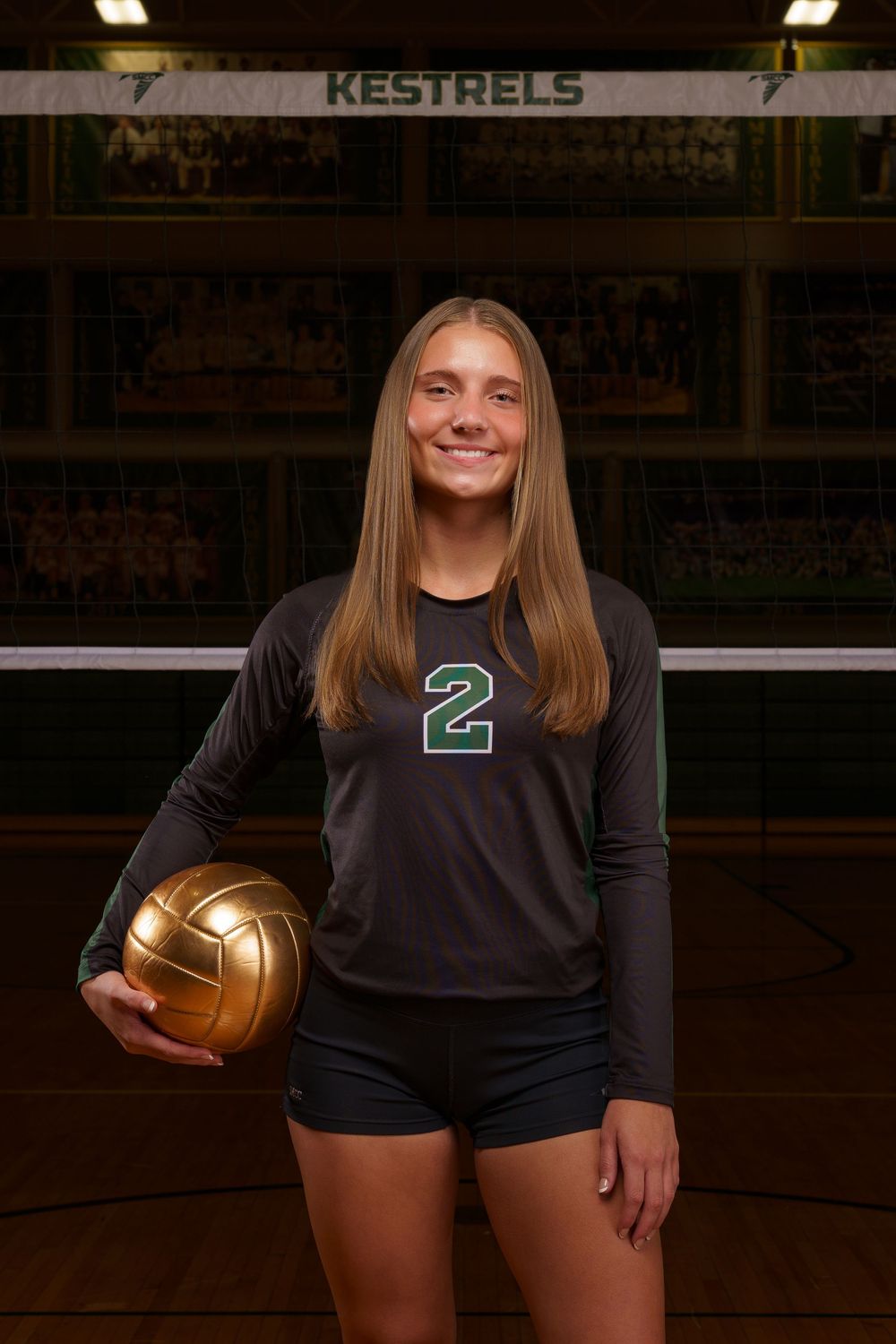 Smiling female volleyball player wearing number 2 jersey holds gold ball in front of Kestrels team banner in gym.