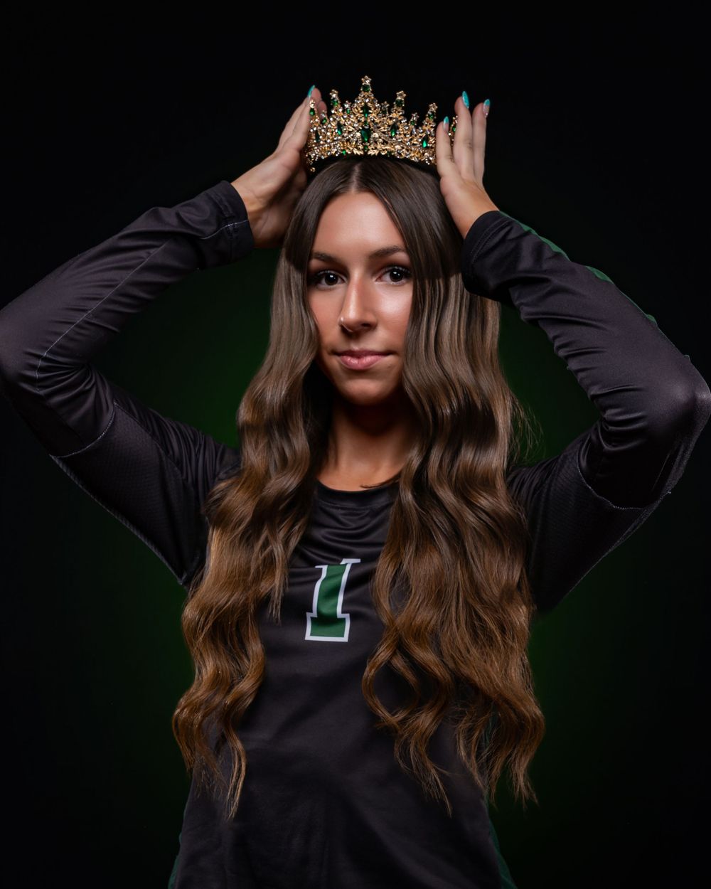 Young woman in black volleyball jersey wearing a gold crown, posing against a dark green background.