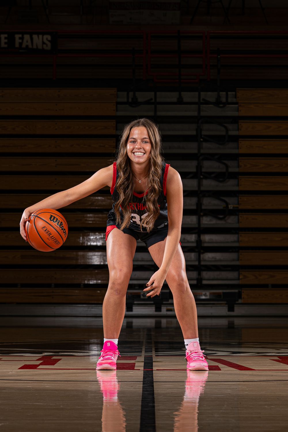 Female basketball player in red jersey smiles while dribbling a Wilson basketball on an indoor court wearing pink sneakers.