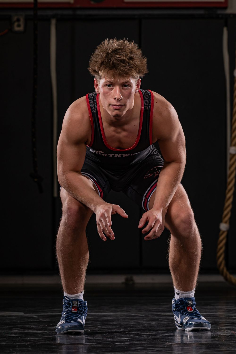 Young male wrestler in black and red singlet crouches in ready stance on dark gym floor.