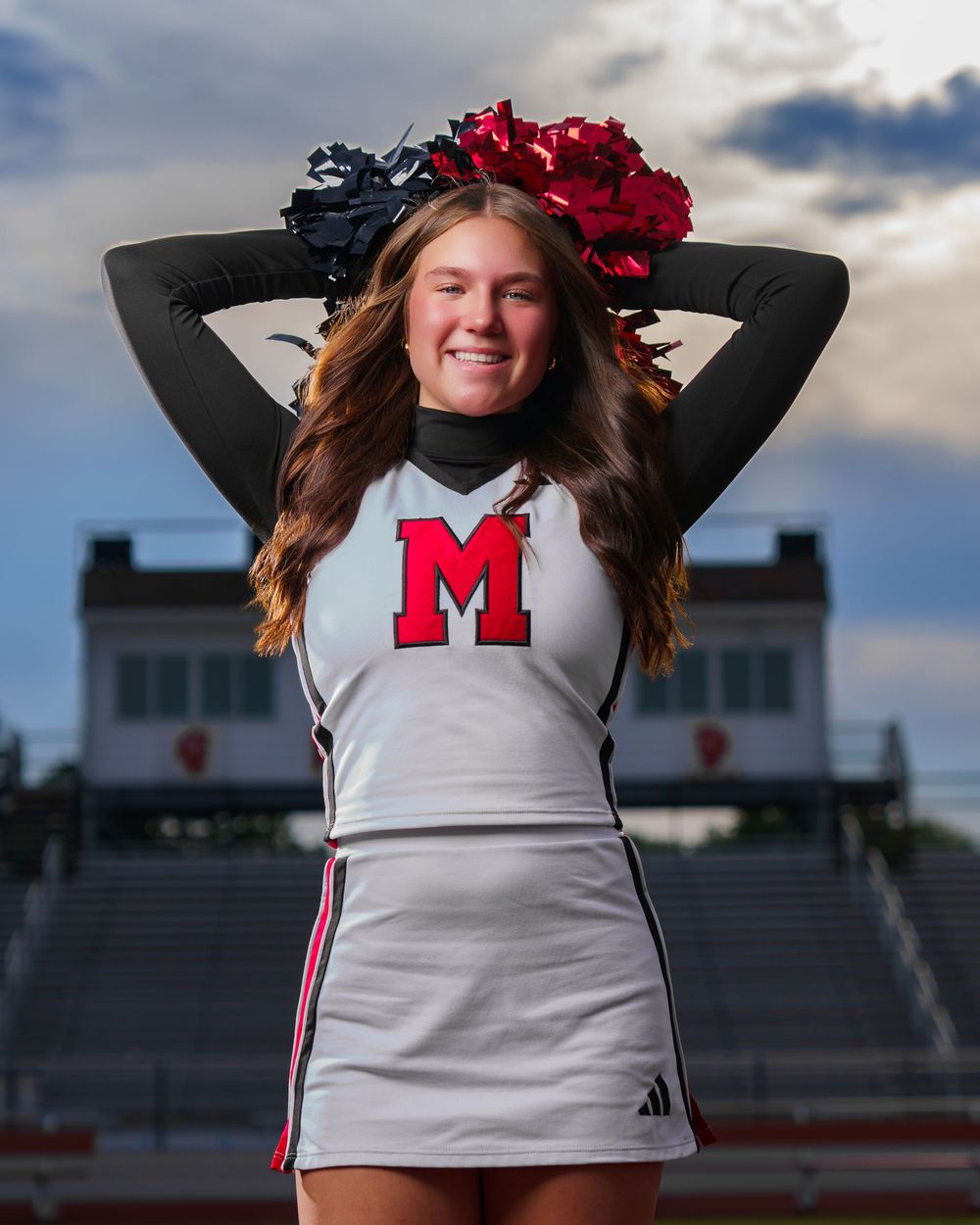 Cheerleader in white uniform with red M logo poses with pom poms at a football stadium.