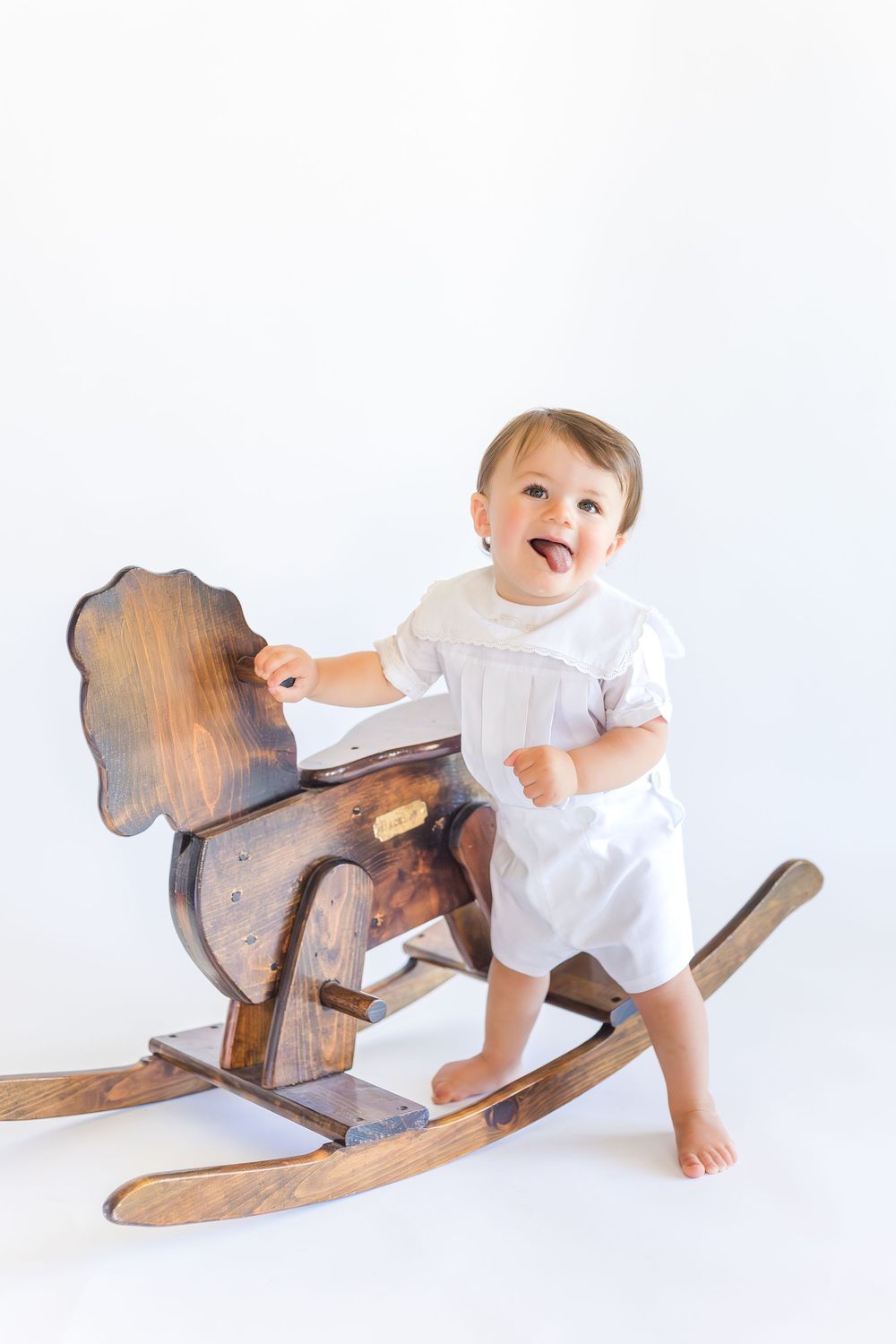 Young child wearing a classic white outfit sitting on a wooden rocking horse against a soft white studio background.