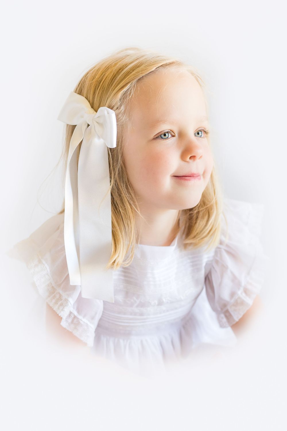 Timeless heirloom portrait of a young blonde girl wearing a white dress and hair bow, looking toward the soft natural light.