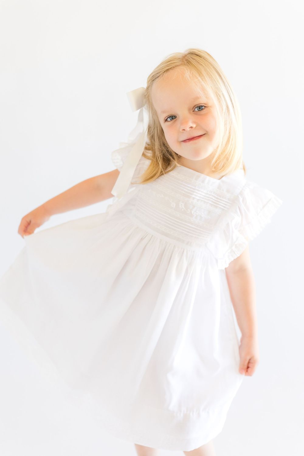 Young blonde girl smiling in a flowing white dress against a bright white background during a classic children's portrait session.