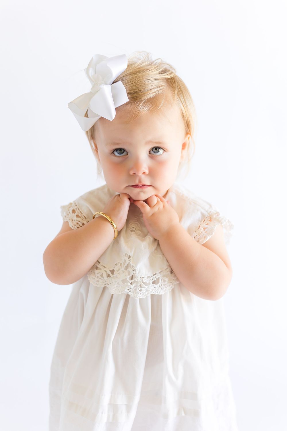 Little girl wearing a white heirloom dress and matching hair bow, holding her hands near her face during a timeless studio portrait.
