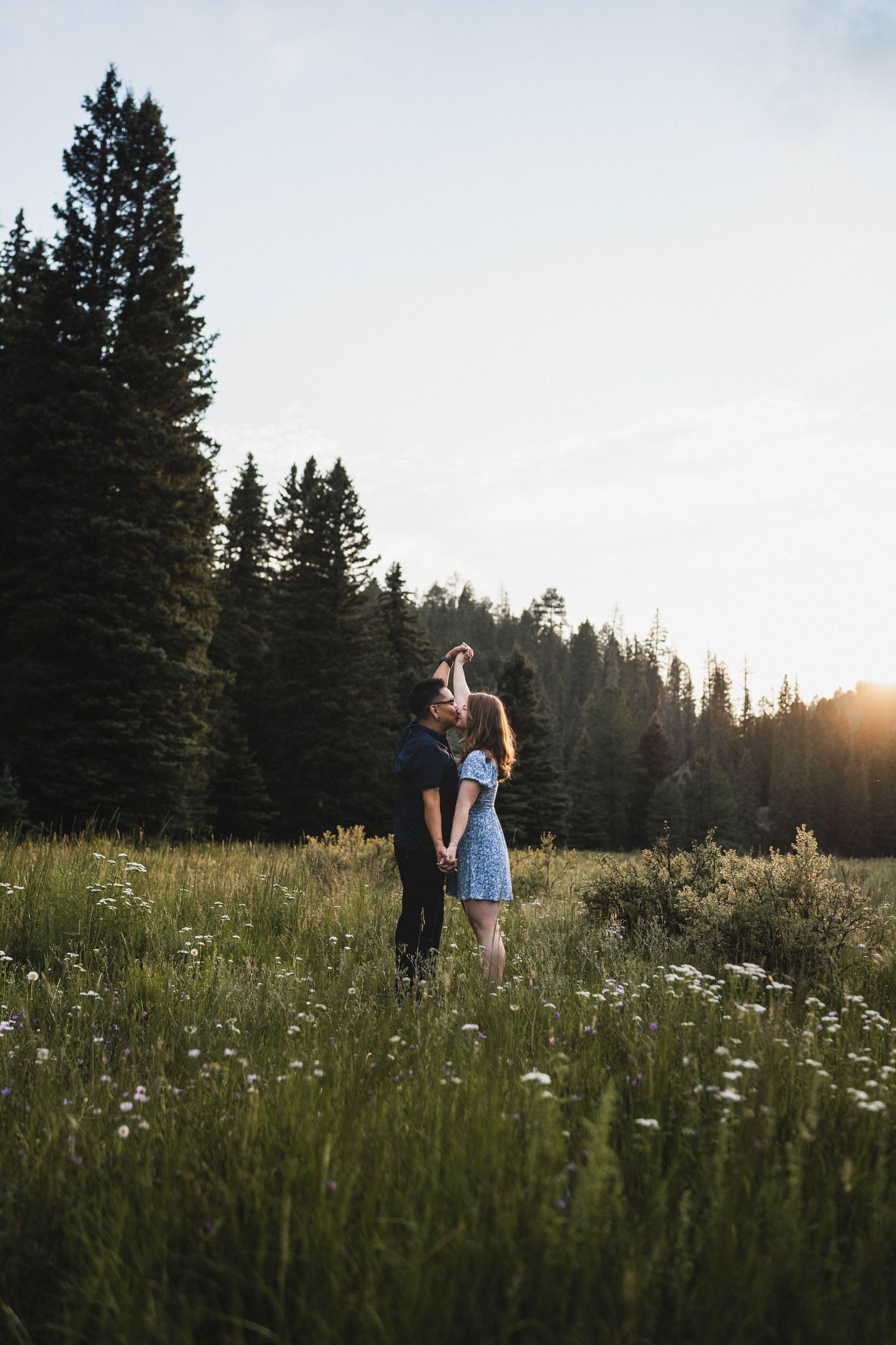 Couple dancing in a wildflower meadow surrounded by tall pine trees at golden sunset.
