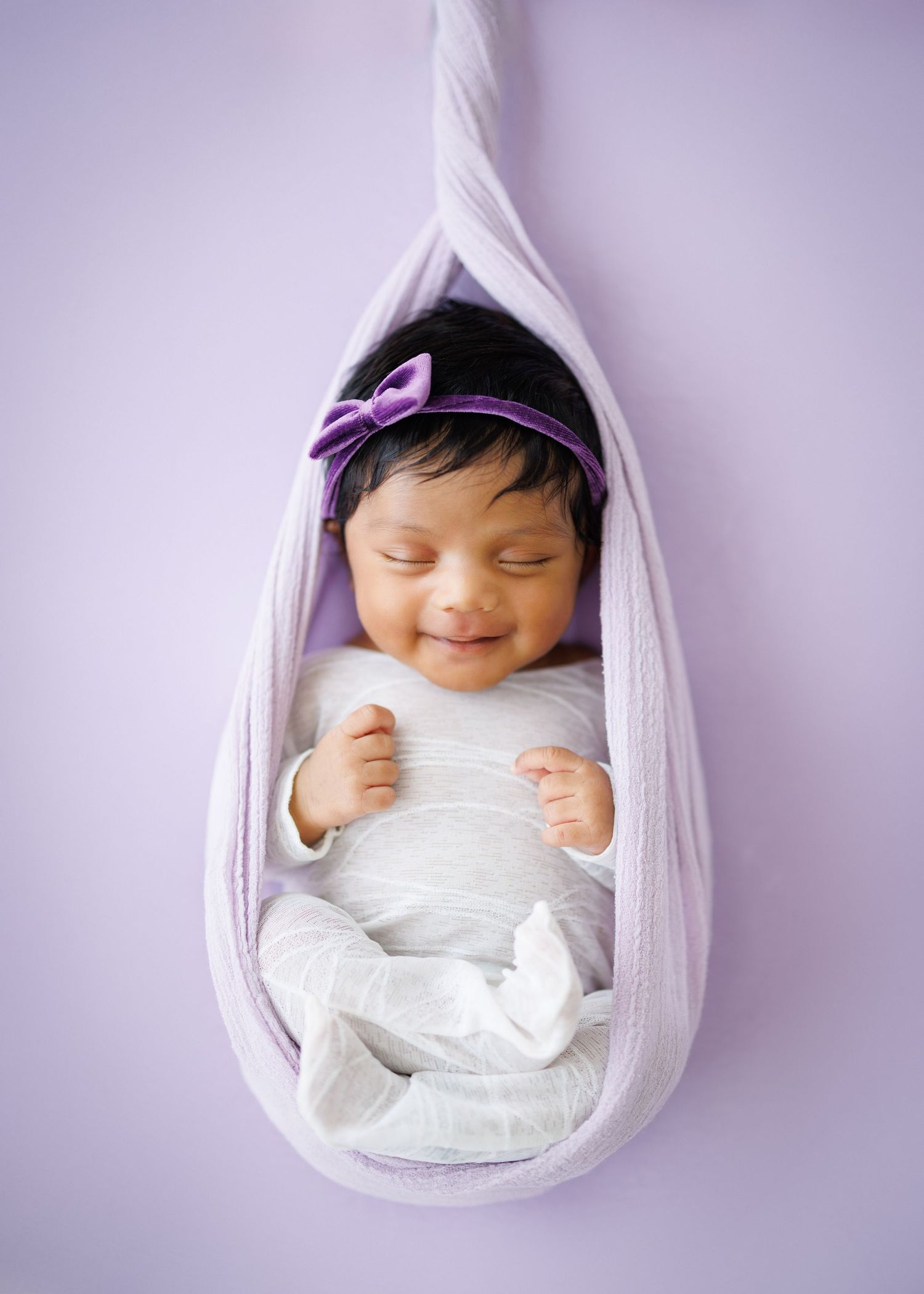 Smiling newborn baby girl wrapped in lavender cloth with purple bow headband, posed in hanging swaddle prop.