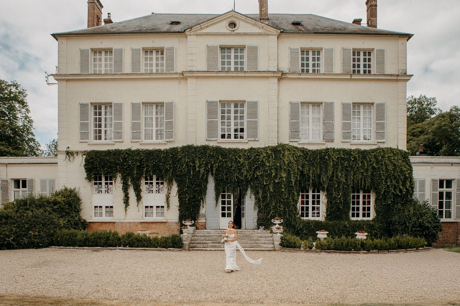 Bride posing in front of an elegant ivy-covered French chateau with white shutters and manicured grounds.