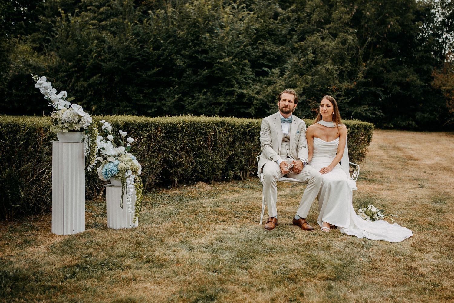 Bride and groom sitting together outdoors on grass, with white floral pedestals and lush green hedges backdrop.
