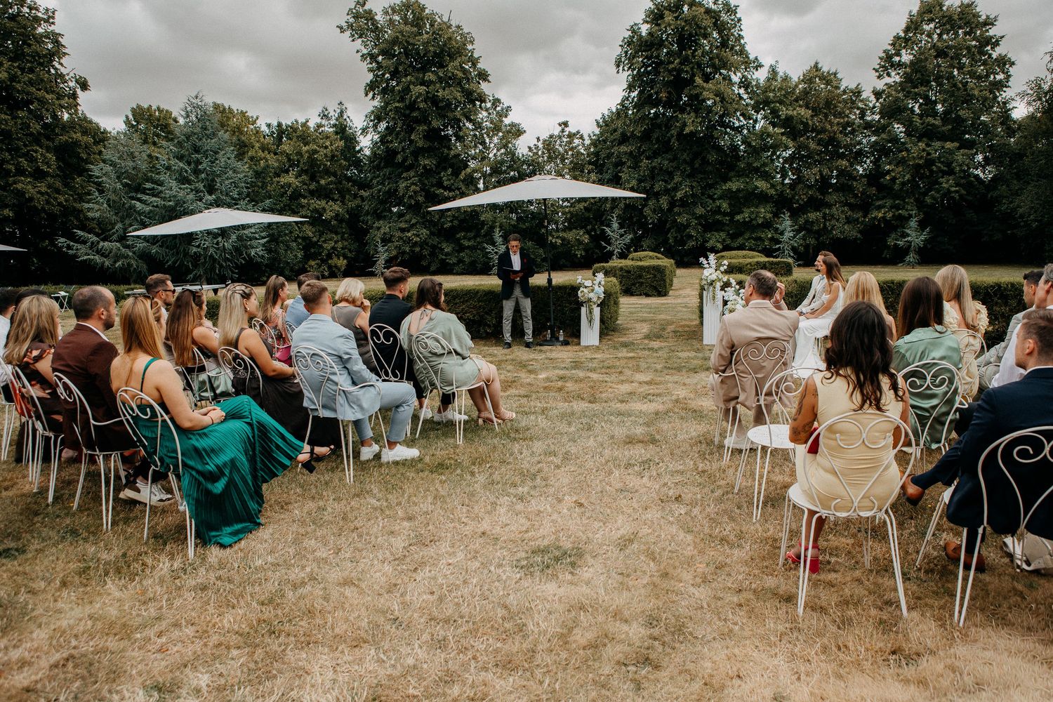 Outdoor wedding ceremony with guests seated on white chairs in a garden setting under cloudy skies.