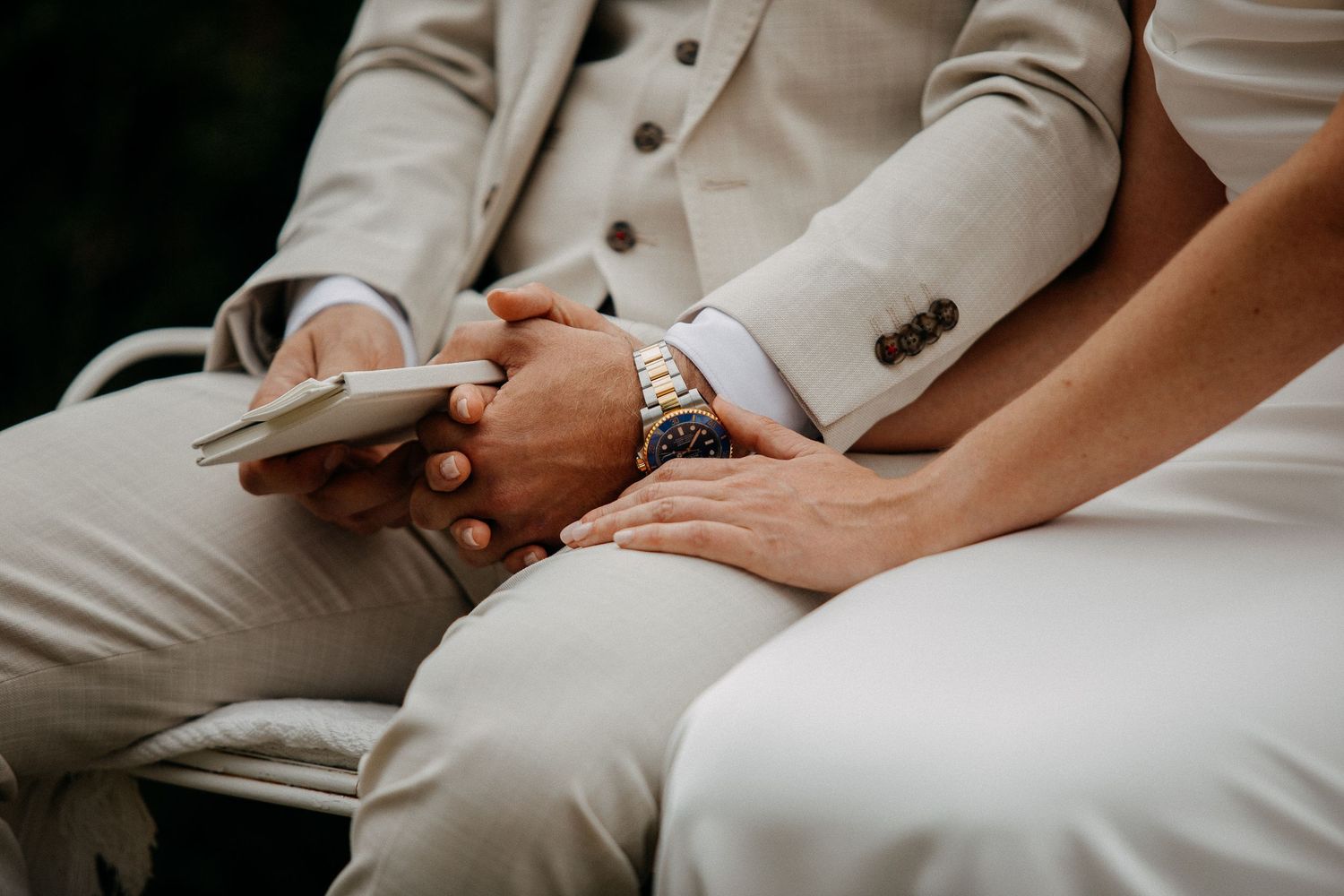 A groom in a beige suit holds a phone while a bride in white gently rests her hand on his, wearing a gold watch.