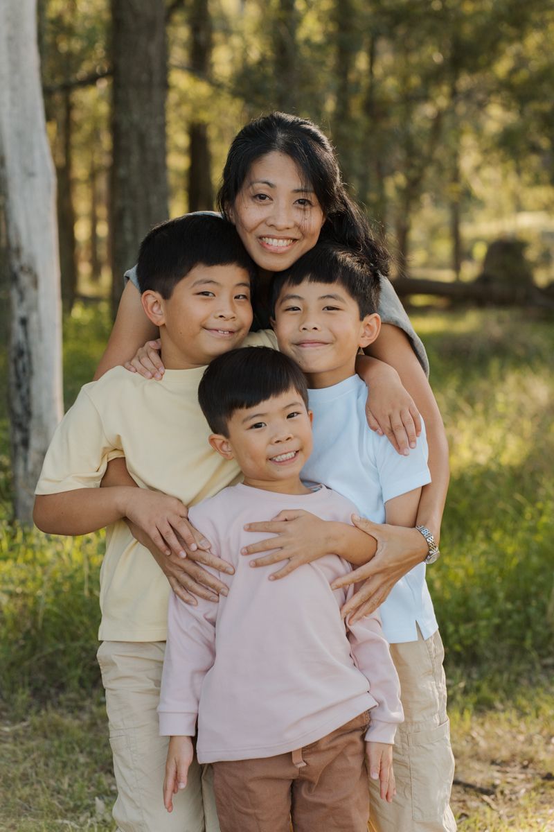 A smiling Asian mother hugs her three young sons outdoors in a sunlit forest setting rouse hill photoshoot