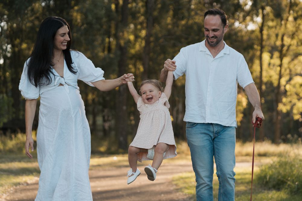 Happy family with pregnant mother in white dress and father swinging toddler girl between them on sunny park path rouse hill family photosho
