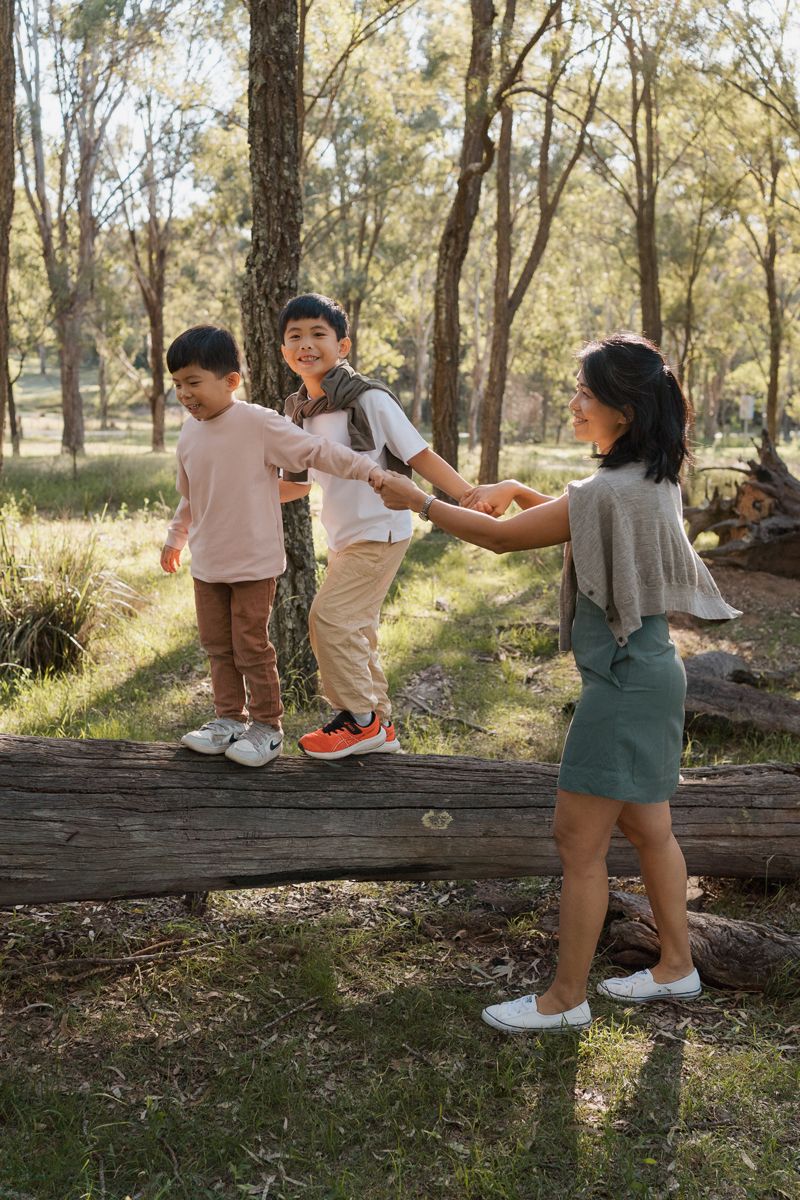 Children playing on a fallen log in a sunny forest park, with a girl helping balance two boys outdoors rouse hill family photoshoot