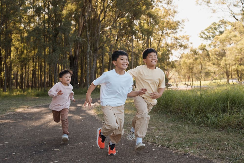 Three children running joyfully on a dirt path through a sunny park surrounded by tall green trees rouse hill family photoshoot