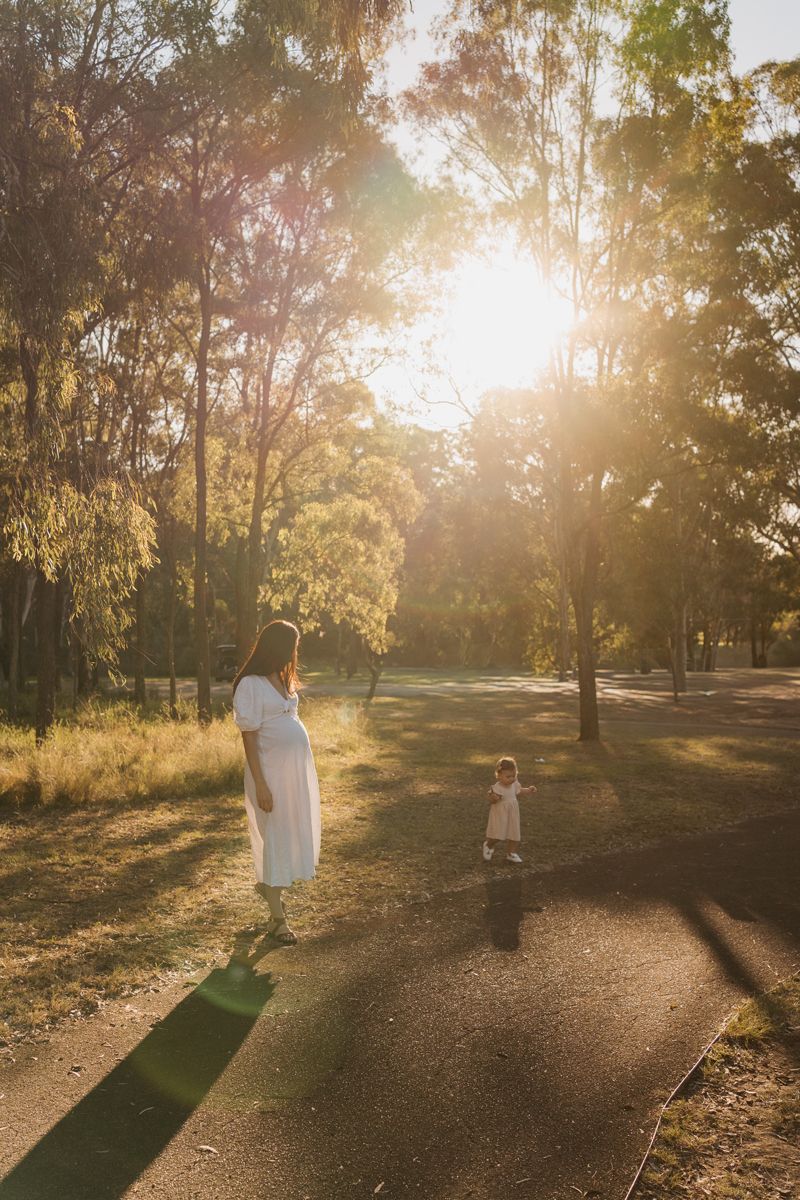 Pregnant woman in white dress walks with toddler on sunlit park path surrounded by tall trees at golden hour Rouse hill family photoshoot