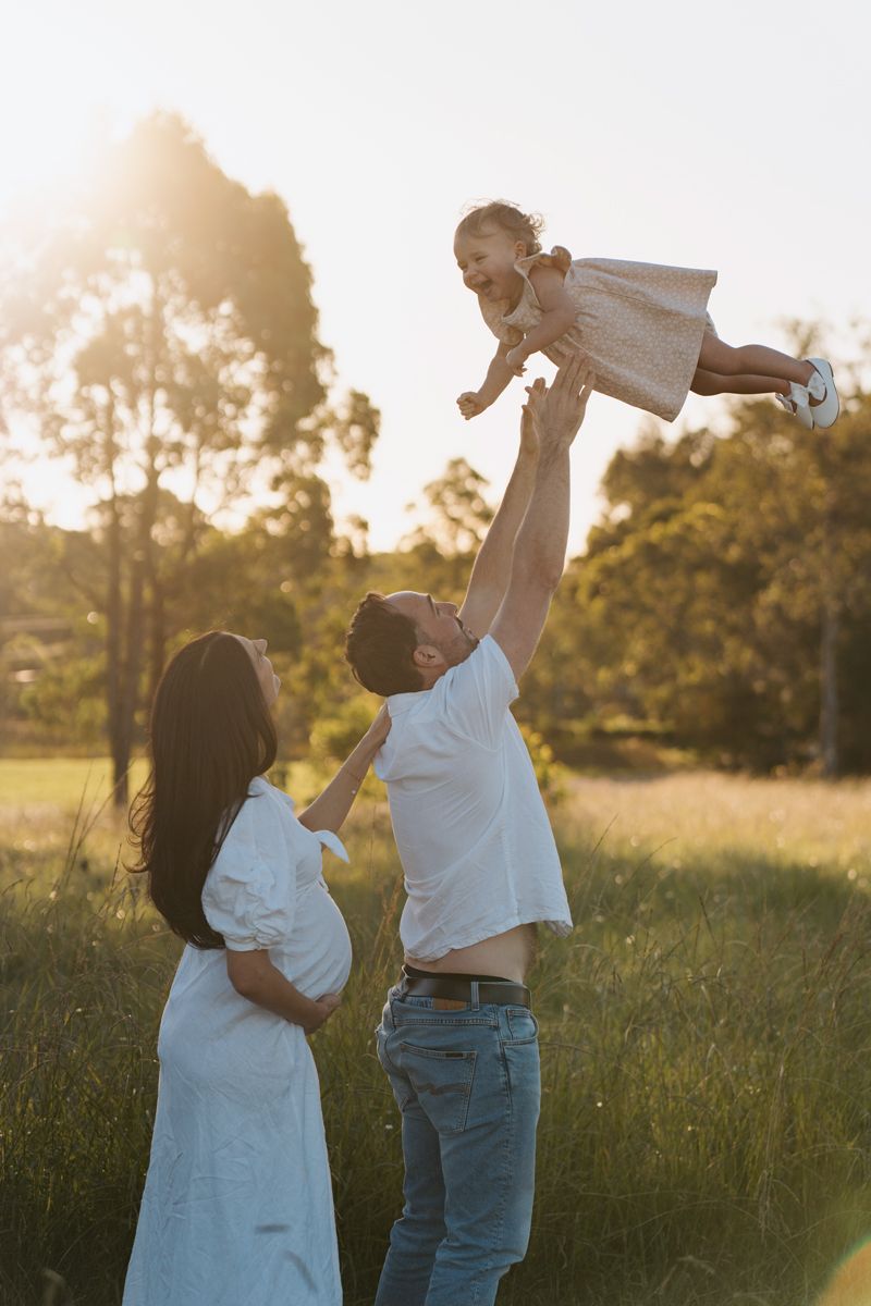 A father lifts his laughing toddler in the air while a pregnant mother watches in a sunlit green field Rouse hill family photoshoot
