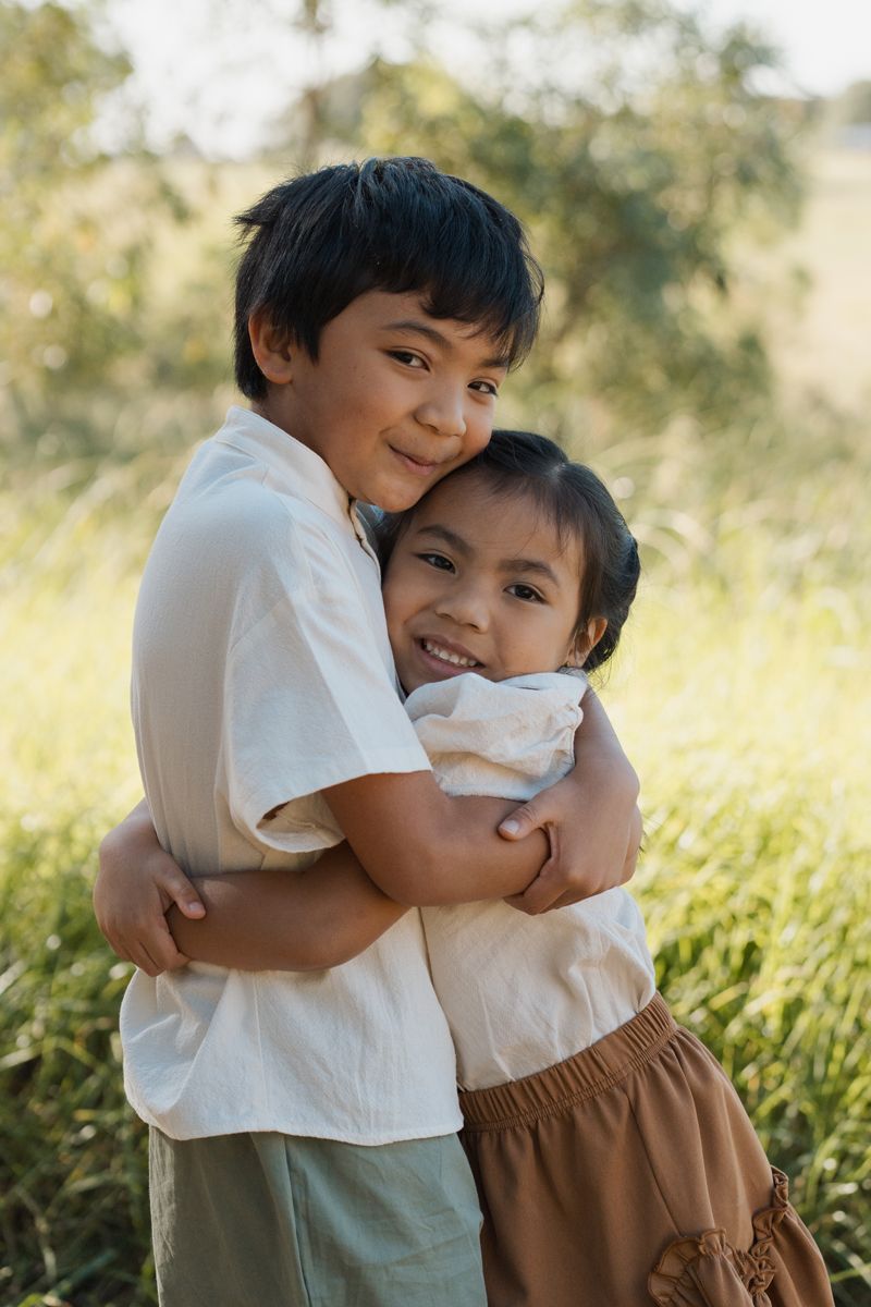 Two young children hugging and smiling outdoors in a green field, wearing casual clothing on a sunny day Rouse hill family photoshoot
