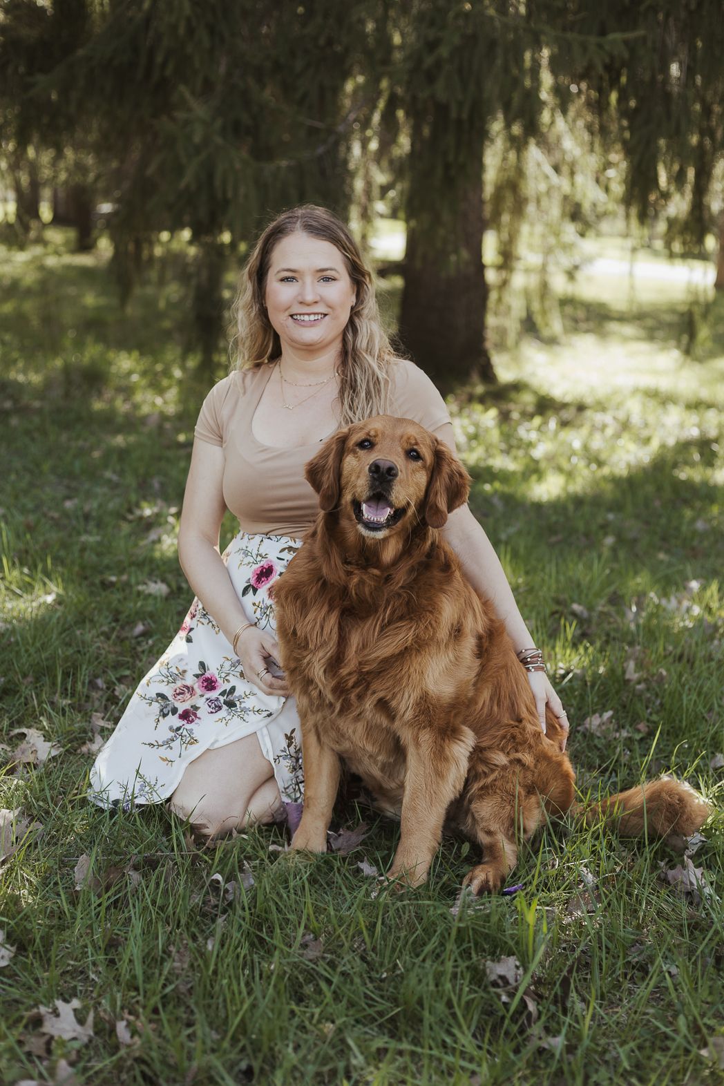 A smiling young woman sits on green grass with a golden retriever dog in a sunlit park setting.