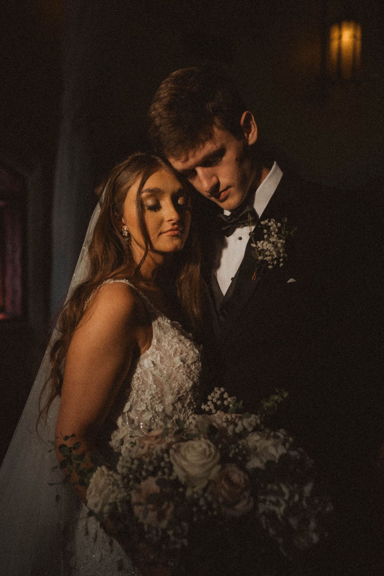 Romantic bride and groom portrait in dramatic moody lighting, bride holding white rose bouquet wearing lace gown with veil.