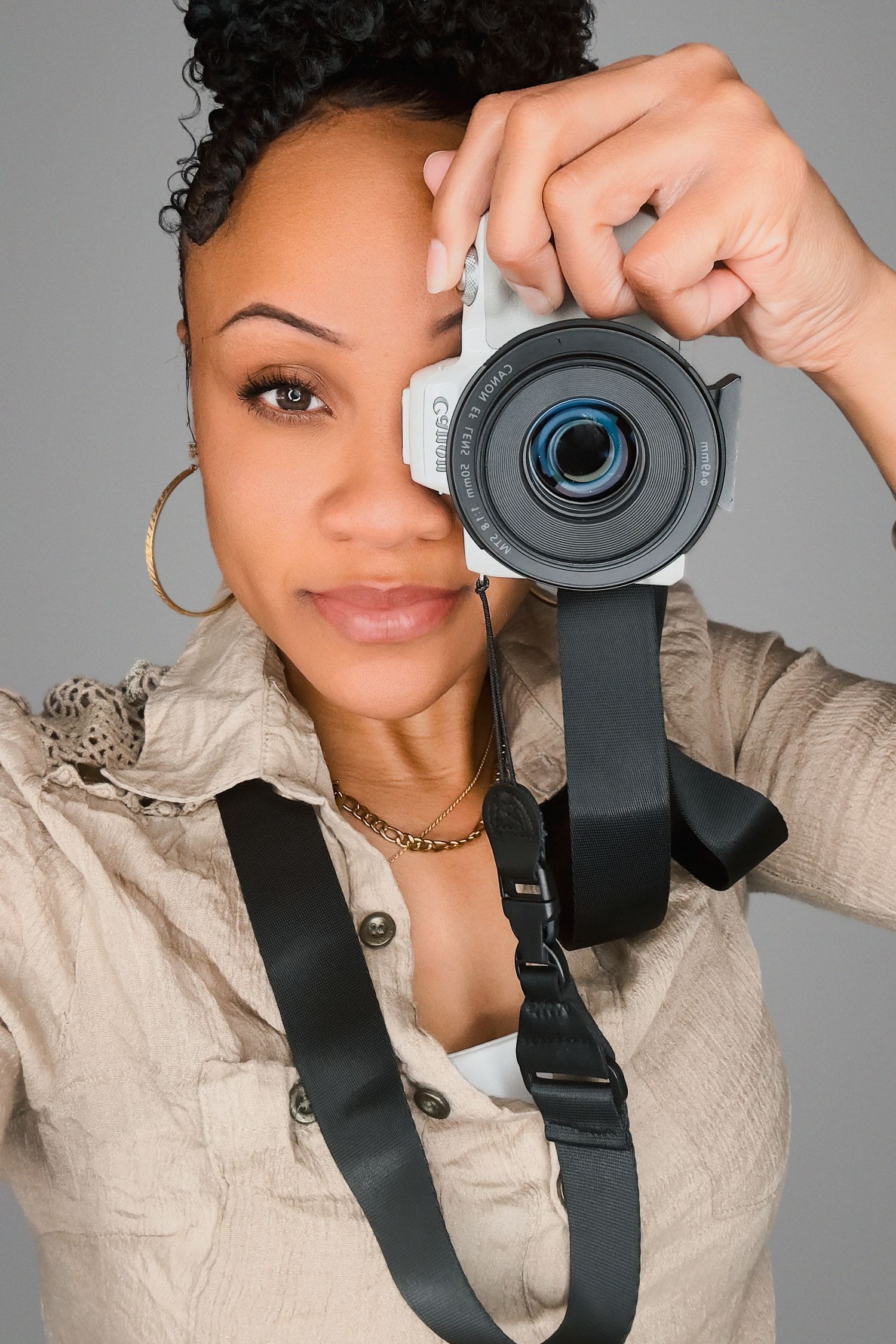 A photographer holds up a digital camera with a black strap while wearing a beige button-up top and gold hoop earrings.