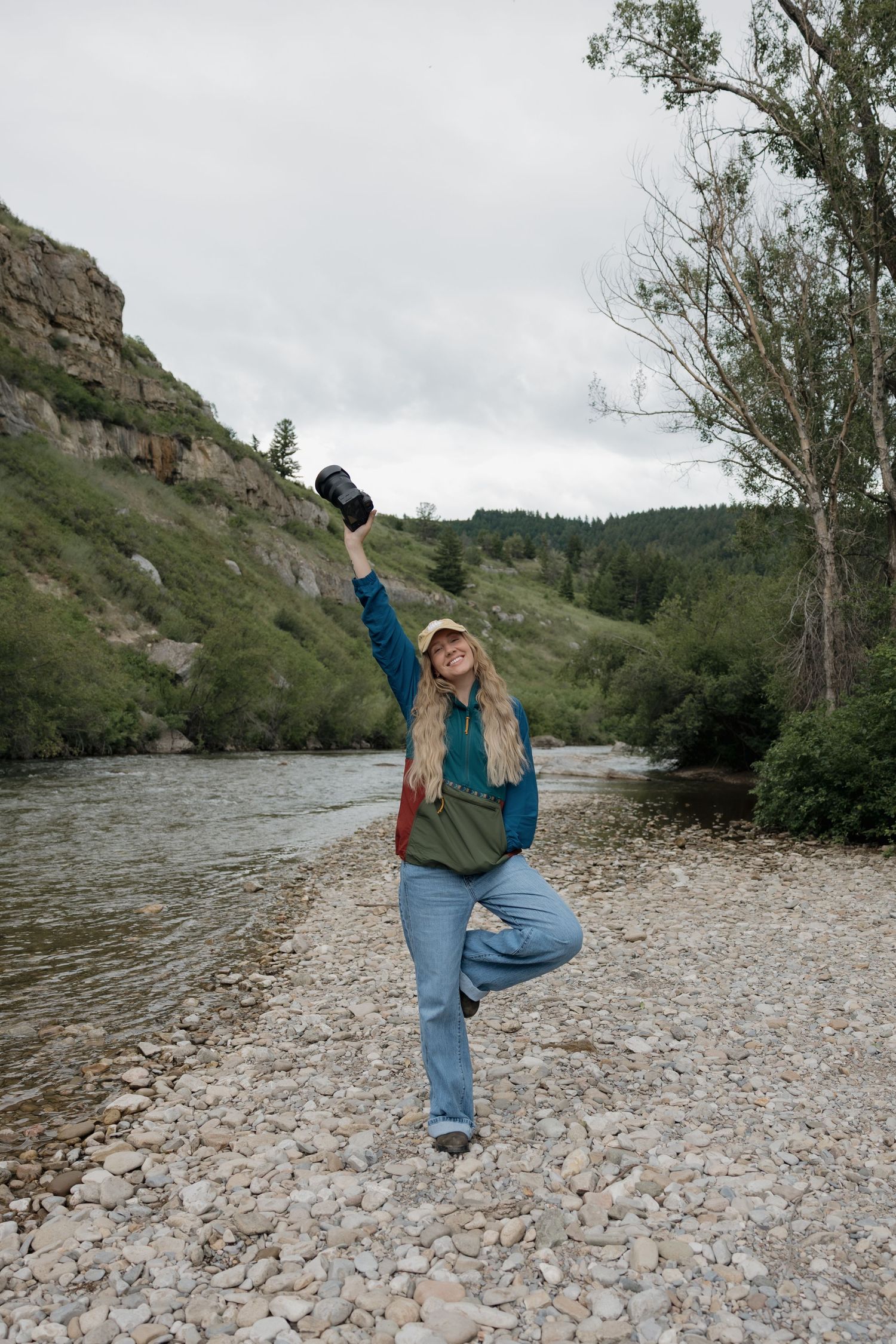 Woman balancing on one leg holding camera by rocky riverside with green hills and trees in background.