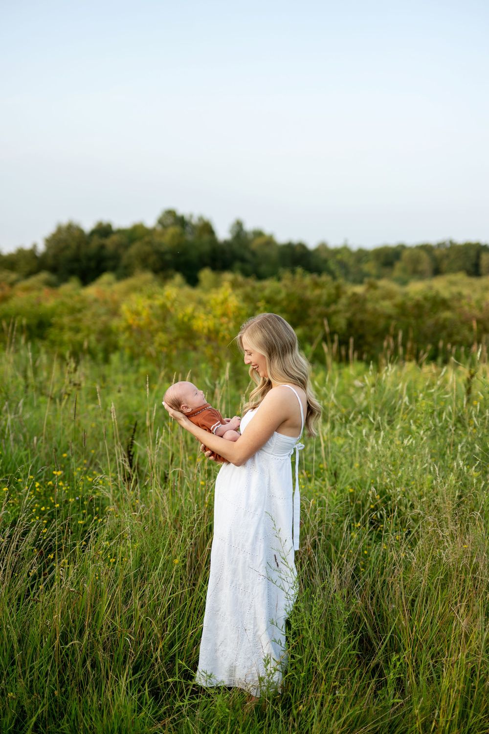 A woman in a white dress holds a newborn baby in a lush green meadow during golden hour.