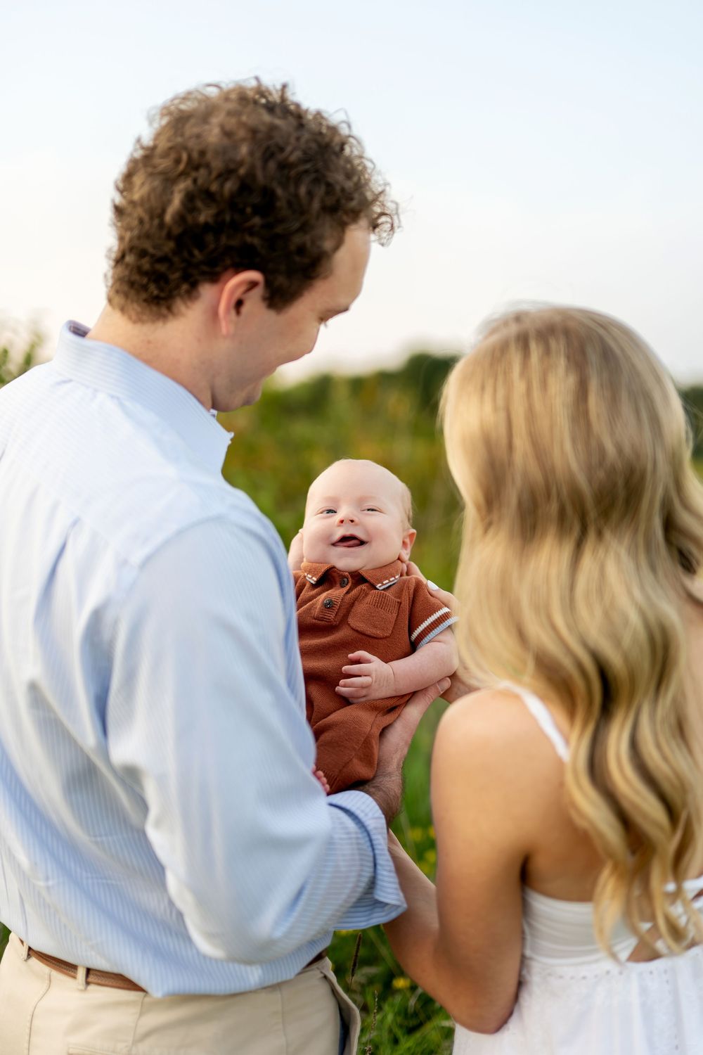 A couple holds their smiling baby outdoors in a green field during golden hour, parents dressed in light blue and white.