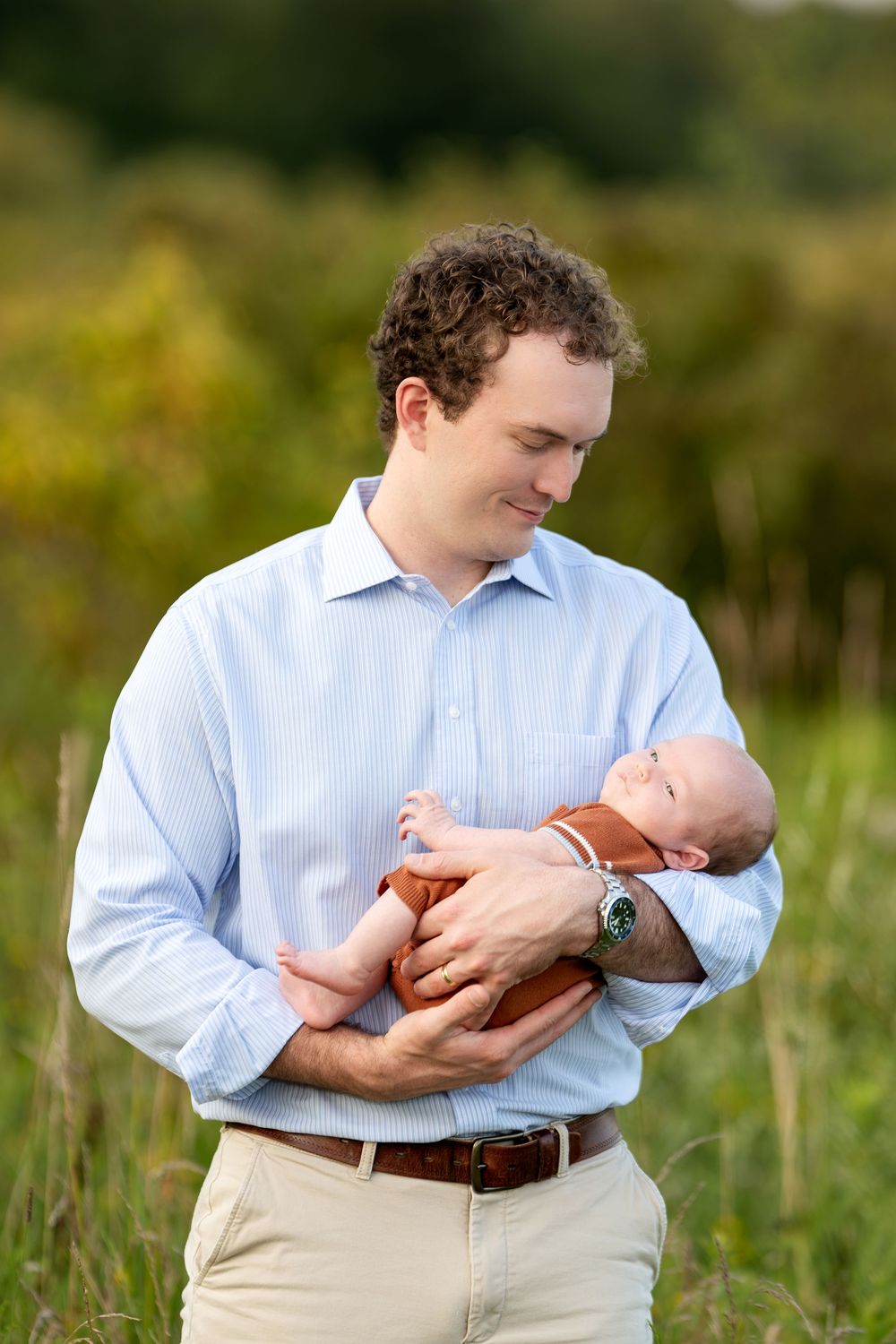 Father in blue shirt tenderly holds newborn baby outdoors in a lush green field during golden hour.