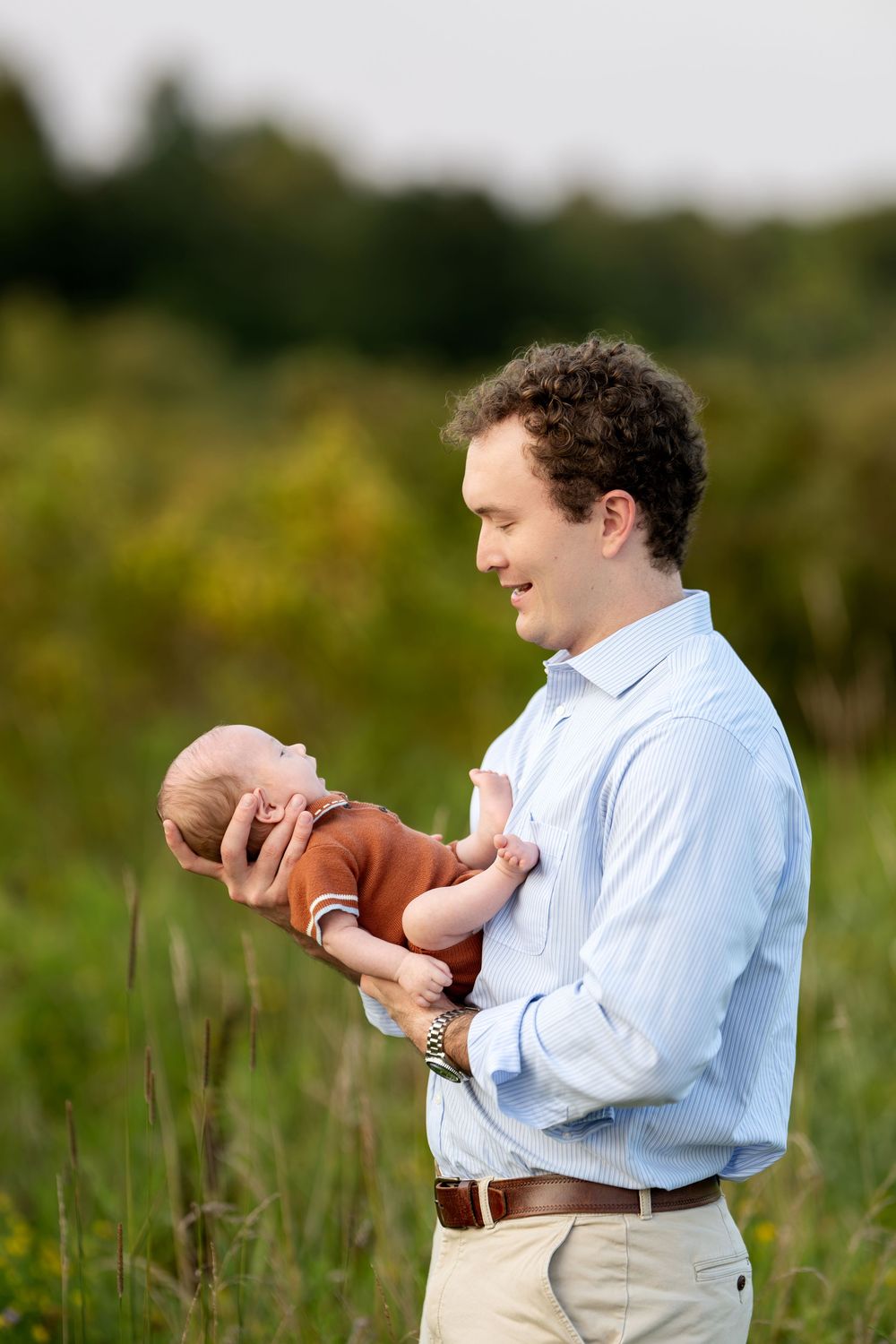 Father smiling while carefully holding his newborn baby in a lush green field outdoors.