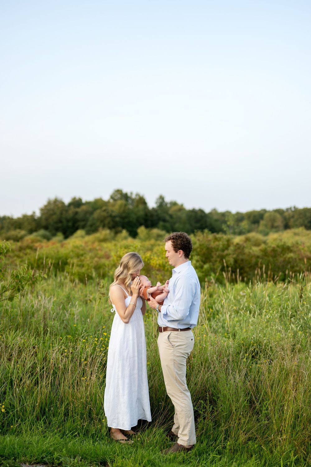 A couple stands in a lush green field holding their newborn baby, dressed in white and blue on a clear day.