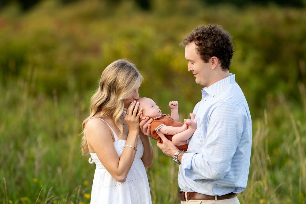 A couple stands in a sunlit meadow, lovingly holding their newborn baby wrapped in an orange blanket.