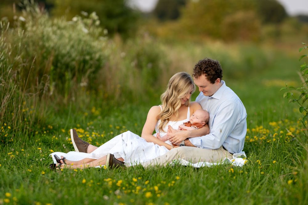 Young couple sitting in green meadow with wildflowers, tenderly cradling their newborn baby together.
