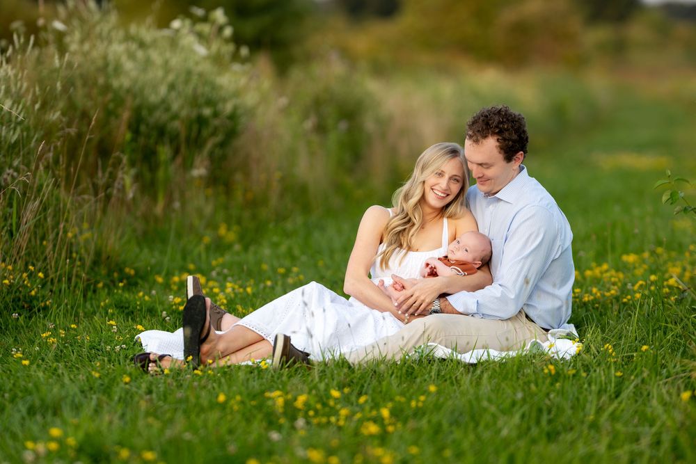 Young family with newborn baby sitting in green meadow with wildflowers during outdoor photo session.