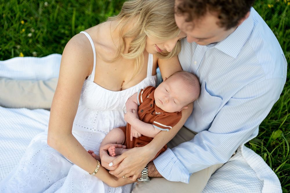 Parents sitting on a blanket in green grass, lovingly cradling their sleeping newborn baby wrapped in a rust-colored swaddle.